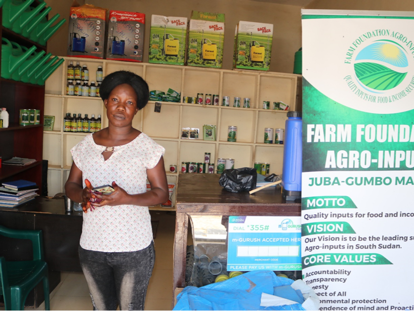 Clara Keji, in a white shirt and blue jeans, stands in her agro-input shop next to a large pop-up banner