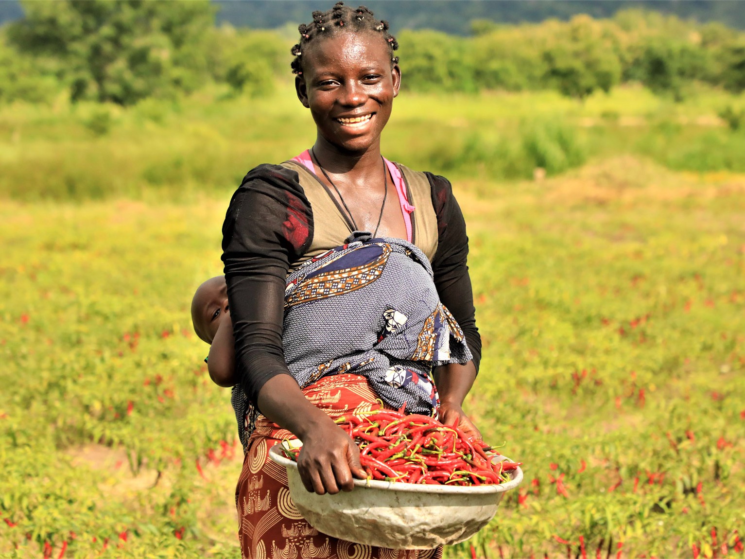 A mother from Benin smiles holding a basket of chili peppers