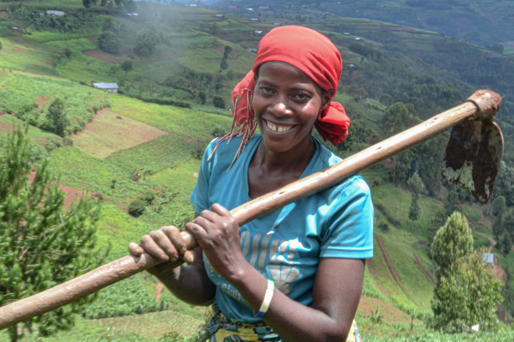 Female farmer with red head covering and blue shirt stands in a field in Uganda